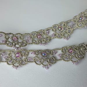 May include: A silver-toned choker necklace with intricate lace-like detailing. The necklace is adorned with small, light purple beads and larger, iridescent pink gemstones. A silver clasp secures the necklace.
