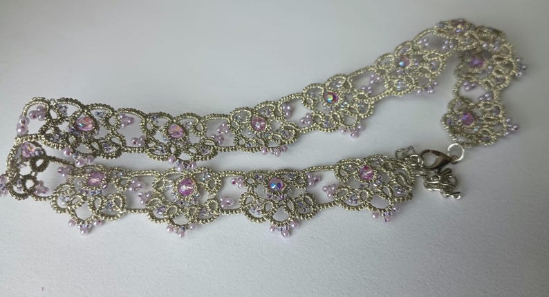 May include: A silver-toned choker necklace with intricate lace-like detailing. The necklace is adorned with small, light purple beads and larger, iridescent pink gemstones. A silver clasp secures the necklace.