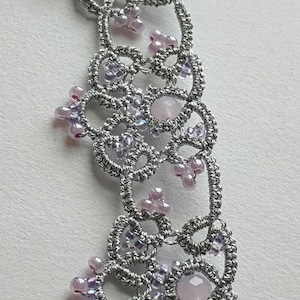 May include: Close-up of a silver-colored, handmade lace necklace. The necklace features a delicate, openwork design with small, pink and purple beads and larger, pale pink gemstones. The intricate pattern is visible.