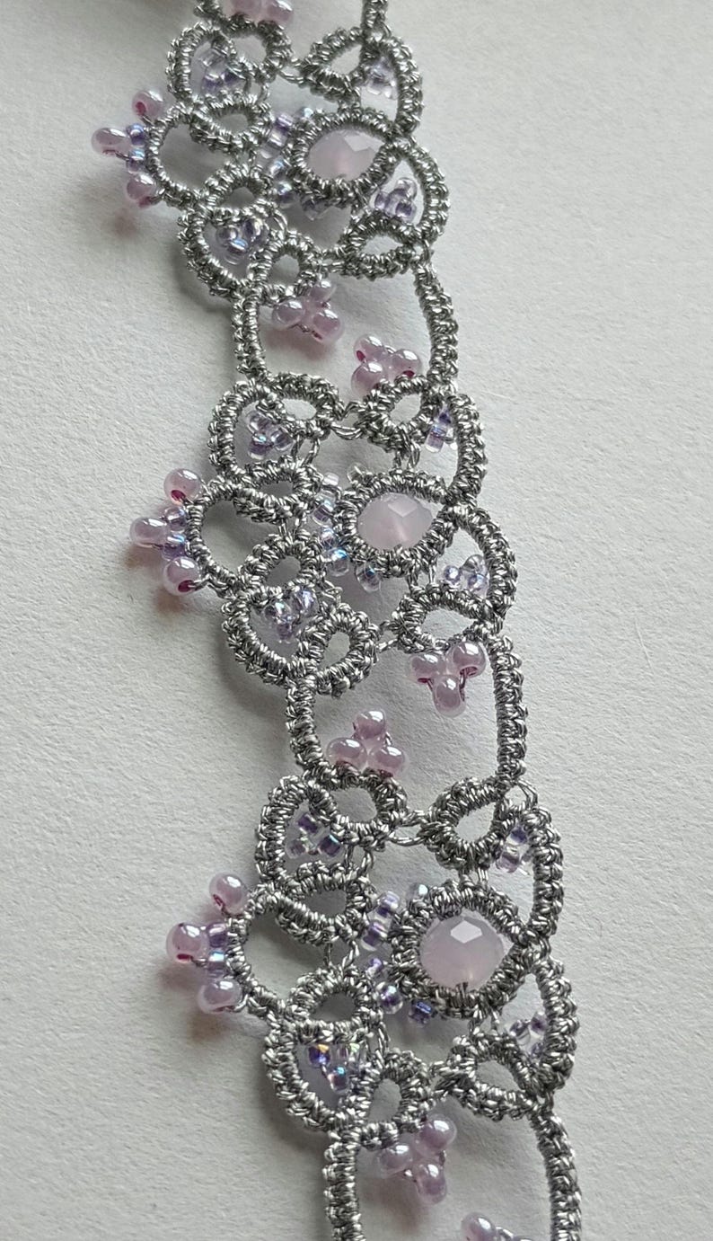 May include: Close-up of a silver-colored, handmade lace necklace. The necklace features a delicate, openwork design with small, pink and purple beads and larger, pale pink gemstones. The intricate pattern is visible.