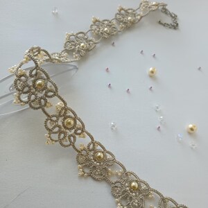 May include: A delicate, silver-toned lace choker necklace with a floral design, adorned with small pearls and larger, golden-colored beads. The necklace is displayed on a clear surface, with scattered pearls and crystals.