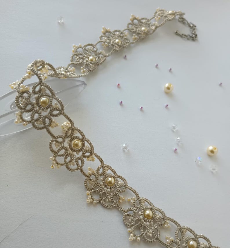 May include: A delicate, silver-toned lace choker necklace with a floral design, adorned with small pearls and larger, golden-colored beads. The necklace is displayed on a clear surface, with scattered pearls and crystals.