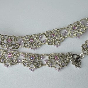 May include: A silver-toned choker necklace with intricate lace-like detailing. The necklace is adorned with small, iridescent pink gemstones and matching beads. A delicate clasp secures the necklace.