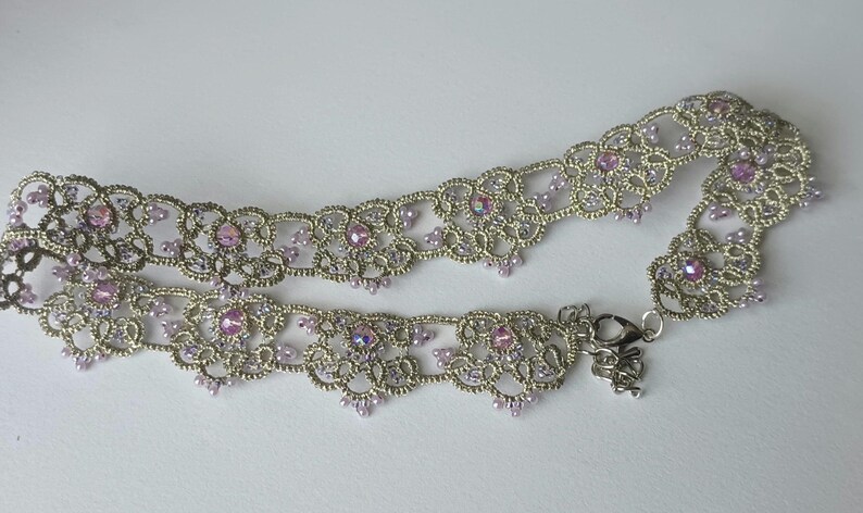 May include: A silver-toned choker necklace with intricate lace-like detailing. The necklace is adorned with small, iridescent pink gemstones and matching beads. A delicate clasp secures the necklace.