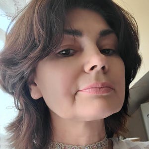 May include: A close-up shot of a person wearing a delicate, ornate choker necklace. The necklace features a light-colored, possibly silver or gold, intricate design with small, pink-toned embellishments. The person has brown hair and is looking towards the camera.