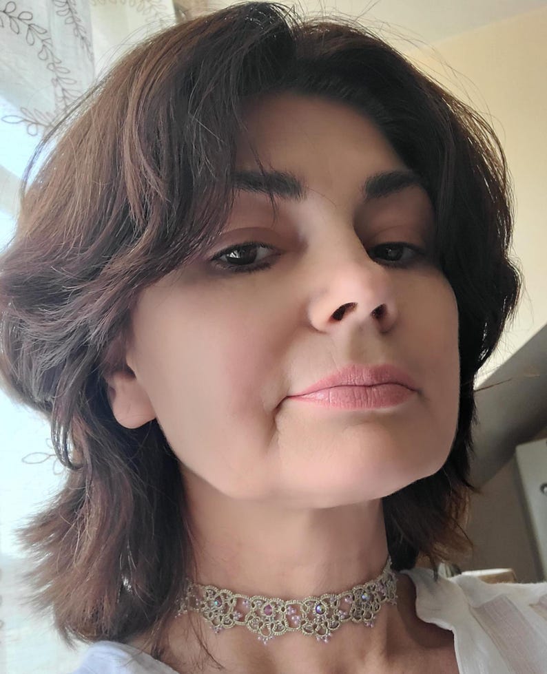 May include: A close-up shot of a person wearing a delicate, ornate choker necklace. The necklace features a light-colored, possibly silver or gold, intricate design with small, pink-toned embellishments. The person has brown hair and is looking towards the camera.