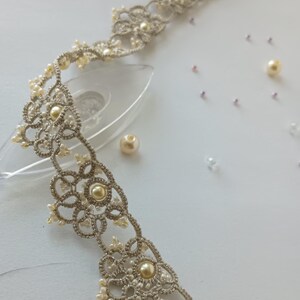May include: A delicate, silver-toned lace choker necklace with pearl accents. The intricate design features floral patterns and small, cream-colored pearls. Scattered pearls and beads add to the elegant aesthetic. The necklace is displayed on a white surface.