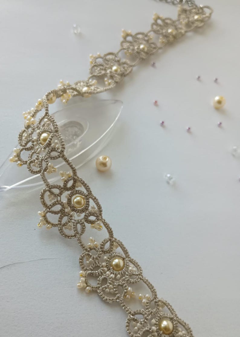 May include: A delicate, silver-toned lace choker necklace with pearl accents. The intricate design features floral patterns and small, cream-colored pearls. Scattered pearls and beads add to the elegant aesthetic. The necklace is displayed on a white surface.