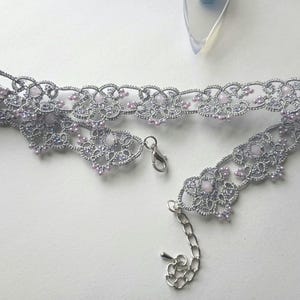 May include: A silver-colored choker necklace with a delicate, lacy design. The necklace features small, light purple beads and a silver clasp. The intricate pattern and color palette create an elegant accessory.