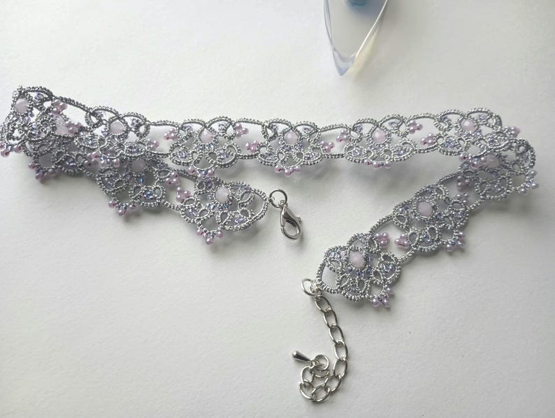May include: A silver-colored choker necklace with a delicate, lacy design. The necklace features small, light purple beads and a silver clasp. The intricate pattern and color palette create an elegant accessory.