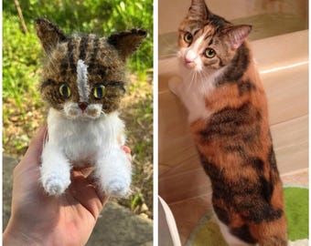May include: A handmade, miniature cat plushie with brown and white fur, held in a hand. The cat has large, expressive eyes and a white stripe down its face. Next to it is a calico cat with orange, brown, and white fur, standing in a bathroom.
