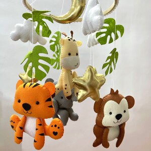 Puede incluir: Un móvil para bebé con temática de la selva. Presenta un tigre, una jirafa, un mono y una estrella. El móvil está hecho de fieltro y tiene un anillo de madera.