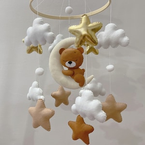 May include: A baby mobile featuring a brown teddy bear resting on a cream-colored crescent moon, surrounded by white clouds, gold and tan stars, and white pom-poms. The mobile is suspended from a wooden hoop.