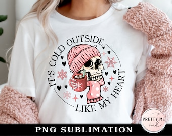 May include: White long-sleeve shirt with a pink and black skull wearing a pink hat and holding a cup of hot chocolate. The text "It's cold outside like my heart" is printed on the shirt.