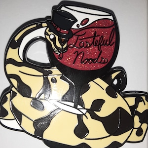 Peut inclure: Un serpent noir et blanc avec un verre de vin rouge sur le dessus. Le verre à vin a une bordure noire et un intérieur blanc. Le verre à vin est rempli de paillettes rouges et le texte "Tasteful Noods" est écrit sur le côté. Le serpent porte un chapeau haut de forme noir avec un ruban rouge.