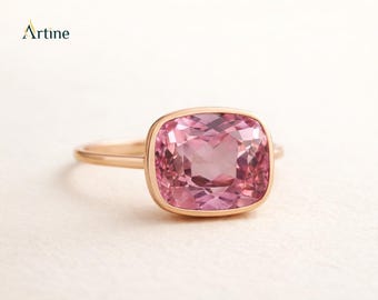 May include: A rose gold ring featuring a large, cushion-cut pink gemstone set in a smooth bezel setting. The ring has a thin, polished band. The word Artine is printed in the upper left corner of the image in black and gold lettering.