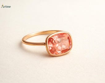May include: A rose gold ring featuring a cushion-cut, faceted peach-colored gemstone set in a smooth bezel. The band is thin and polished, with the brand name Artine printed in the upper left corner of the frame. The design is simple and elegant.