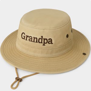 May include: Tan bucket hat with a wide brim and a chin strap. The word "Grandpa" is embroidered in brown on the front of the hat. The hat has a round crown and a slightly sloped brim, with a drawstring.