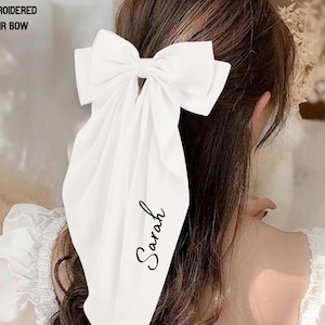 May include: A white embroidered hair bow featuring a large bow and long, flowing fabric. The name "Sarah" is written in cursive on the fabric. The text "EMBROIDERED HAIR BOW" is at the top left.