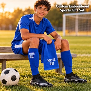 May include: A blue sports uniform set including a short-sleeved jersey, shorts, and blue socks with white text reading MILLER #21. A blue towel with the same white text is included. A soccer ball sits on the grass nearby.