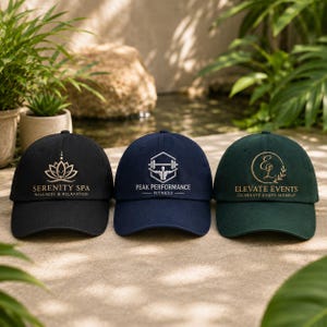 Pode incluir: Três bonés de beisebol nas cores preto, azul marinho e verde escuro. O boné preto apresenta um logotipo de lótus dourado e o texto Serenity Spa Wellness & Relaxation. O boné azul marinho tem um logotipo de barra branca e o texto Peak Performance Fitness. O boné verde exibe um logotipo circular dourado com EL e o texto Elevate Events Celebrate Every Moment.