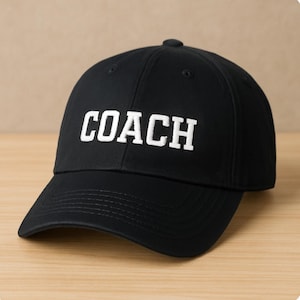 Könnte beinhalten: Schwarze Baseballkappe mit dem Wort "COACH" in weißen Blockbuchstaben auf der Vorderseite gestickt. Die Kappe hat einen gebogenen Schirm und einen Knopf oben. Der Hintergrund ist eine hellbraune Holzoberfläche.