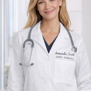 May include: A person wearing a white lab coat and a stethoscope. The lab coat has the name "Amanda Scott" and "APRN - Pediatrics" embroidered on the chest. The background is a blurred interior with a window.