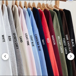 May include: A collection of crew neck t-shirts hanging on wooden hangers, displayed in a row. The colors include white, ash, sports grey, sand, light pink, light blue, carolina blue, navy, red, maroon, military green, forest green, dark chocolate, and black.