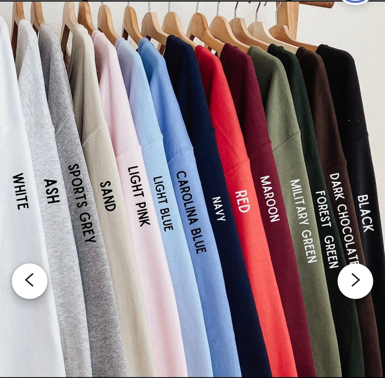 May include: A collection of crew neck t-shirts hanging on wooden hangers, displayed in a row. The colors include white, ash, sports grey, sand, light pink, light blue, carolina blue, navy, red, maroon, military green, forest green, dark chocolate, and black.