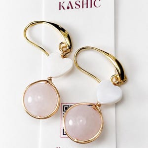 May include: A pair of gold-tone hook earrings featuring a smooth, round, pale pink stone encased in a gold wire frame, suspended below a small white heart-shaped accent. The earrings are displayed on a white card labeled KASHIC HANDCRAFTED.