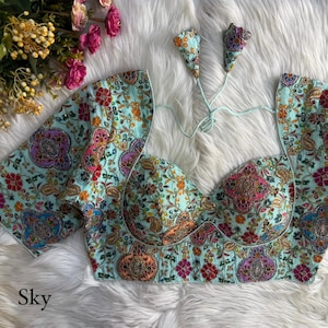 Pode incluir: Top bordado azul celeste com intrincados padrões florais e paisley em tons de rosa, laranja e roxo. O top apresenta mangas curtas e decote em coração. A palavra "Sky" está impressa na parte inferior esquerda.