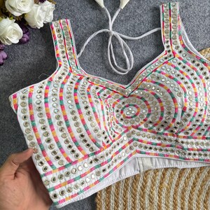 May include: A white sleeveless blouse featuring a square neckline and back tie closure. The garment is decorated with vertical and circular rows of small round mirrors and multi-colored thread embroidery in pink, yellow, and green stripes.