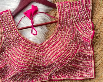 May include: A vibrant fuchsia top with extensive beadwork, featuring a deep V-neck. The design incorporates rows of gold and silver beads, forming a striped pattern. It is displayed on a wooden hanger, highlighting the detailed craftsmanship and vivid colour.