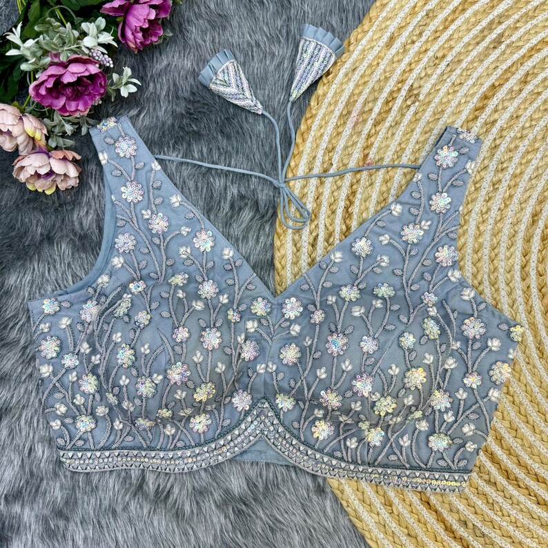 May include: A gray embroidered blouse with a V-neckline and a floral design. The blouse has a tie-up back and is decorated with sequins.