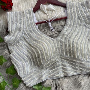 Pu&ograve; includere: Una camicetta bianca senza maniche con intricati motivi verticali e diagonali di paillettes argentate e piccole perle bianche. Il capo presenta uno scollo a V e include lacci in tessuto bianco sul retro per la chiusura.