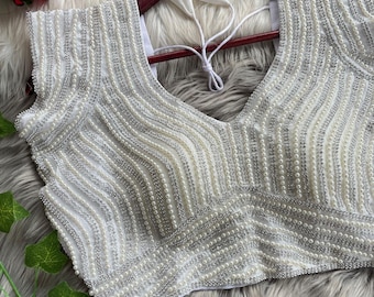 May include: A white sleeveless blouse featuring intricate vertical and diagonal patterns of silver sequins and small white faux pearls. The garment has a V-neckline and includes white fabric ties at the back for closure, creating a textured appearance.