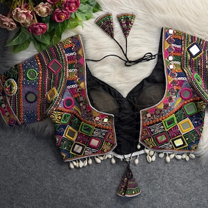 May include: A colorful, embroidered top with a V-neck and short sleeves. The top features a variety of geometric patterns, mirrors, and beadwork in shades of blue, pink, green, and orange. Small white shells adorn the bottom edge. Matching earrings are included.