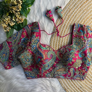 Pode incluir: Uma blusa rosa vibrante com bordados paisley intrincados em tons de azul, verde e dourado. A blusa apresenta um decote quadrado, mangas curtas e fechos de laço. O design ornamentado é definido em um fundo branco e amarelo.