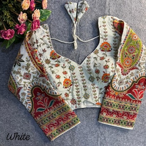 Pode incluir: Blusa branca com bordados florais intrincados em laranja, vermelho e verde. As mangas compridas e o decote são adornados com padrões detalhados. A palavra "White" está impressa na parte inferior esquerda.