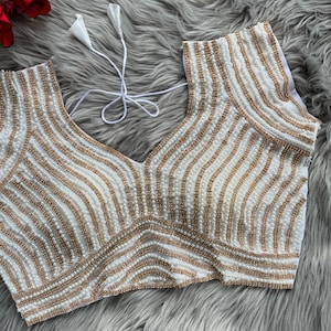 Pu&ograve; includere: Una blusa bianca senza maniche con un profondo scollo a V e intricati motivi a righe verticali realizzati con paillettes dorate e perle artificiali bianche. Il capo presenta lacci in tessuto bianco sul retro del collo e un corpetto aderente.