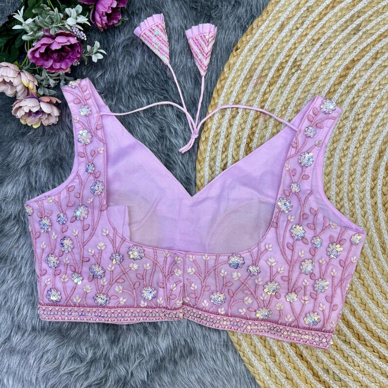 May include: A light purple embroidered bra with a V-neckline and a tie closure in the back. The bra is decorated with sequins and floral patterns.