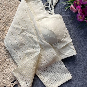 Pode incluir: Blusa marfim bordada com ilhós, com corpete e mangas ajustadas. A blusa apresenta um design delicado e vazado e alças amarradas nas costas. O tecido parece leve e respirável, adequado para climas quentes.
