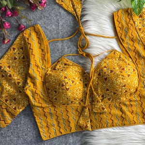 May include: A mustard yellow blouse featuring a sweetheart neckline, long sleeves, and a repeating geometric pattern in shades of brown, white, and blue. The garment includes thin tie-up straps at the shoulders with decorative fabric tassels.