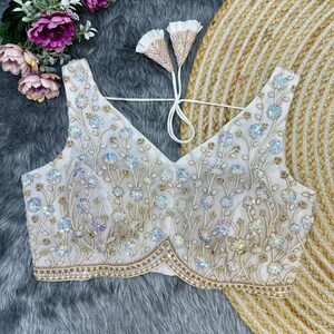 May include: White embroidered blouse with a V-neckline and a floral design. The blouse is decorated with sequins and beads.
