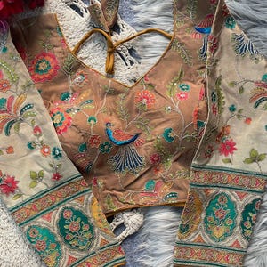 Pode incluir: Uma blusa marrom bordada com mangas compridas. A blusa apresenta um decote em V e é adornada com motivos florais e de pássaros coloridos. As mangas têm uma borda decorativa com um padrão verde e dourado. A blusa é feita de um material macio e sedoso.