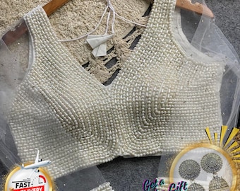 May include: Cream-coloured, beaded top with a deep V-neck and sheer sleeves. The top is adorned with small, round beads, and is displayed on a wooden hanger. The text "The Traditional Street" is visible at the top. A "Fast Delivery" graphic is in the lower left.