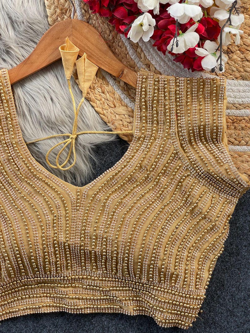 Pu&ograve; includere: Una blusa dorata senza maniche con un profondo scollo a V e intricate file verticali di perline e strass scintillanti. Il capo presenta sottili lacci in tessuto dorato sul retro con estremit&agrave; appuntite e un aspetto strutturato.