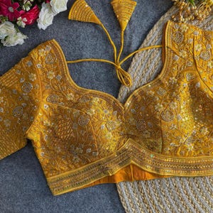 Puede incluir: Una blusa mostaza, bordada intrincadamente, con un escote en V profundo. La blusa presenta patrones florales y geométricos detallados con adornos de lentejuelas. Las mangas son cortas y tiene borlas decorativas.