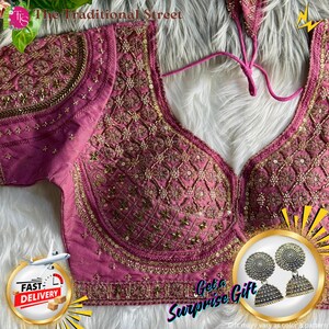 Pode incluir: Uma blusa magenta bordada com detalhes dourados intrincados. A blusa apresenta um decote em V profundo e é adornada com lantejoulas e contas. A imagem também inclui um par de brincos de prata e o texto "The Traditional Street".