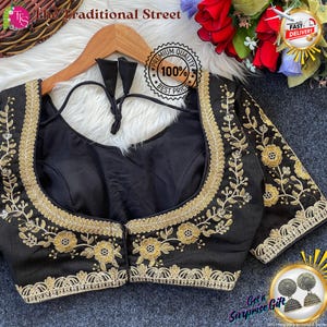 Pode incluir: Blusa preta com bordado floral dourado. A blusa tem decote redondo e mangas curtas. O texto "The Traditional Street" está no topo. A blusa está pendurada em um cabide de madeira.